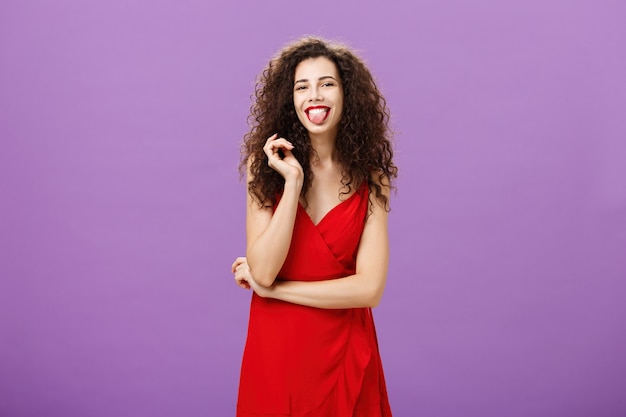 Prise de vue en studio d'une étudiante caucasienne artistique ludique et joyeuse en robe de soirée rouge qui sort la langue joyeusement en s'amusant à jouer avec curl debout sur fond violet insouciant et insouciant.