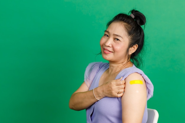 Prise de vue en studio d'une femme asiatique d'âge moyen assise souriante et montrant un bandage en plâtre jaune sur le bras après avoir reçu la vaccination contre le coronavirus covid 19 par un médecin à la clinique sur fond vert.