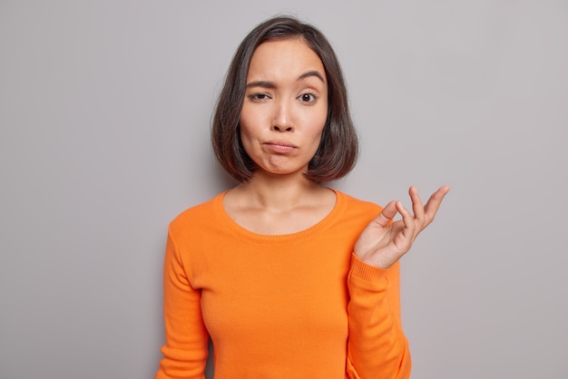 Prise de vue en studio d'une femme asiatique indignée lève la paume avec hésitation ne peut pas prendre de décision vêtue de poses décontractées de cavalier orange contre le mur gris du studio regarde avec perplexité entend quelque chose d'étrange