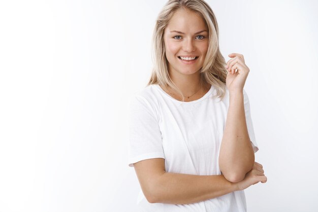 Photo prise de vue en studio d'une femme blonde en bonne santé et séduisante, tatouée sur le bras, souriante, croisée sur le corps, regardant positivement la caméra avec un large sourire amical posant sur fond blanc en riant