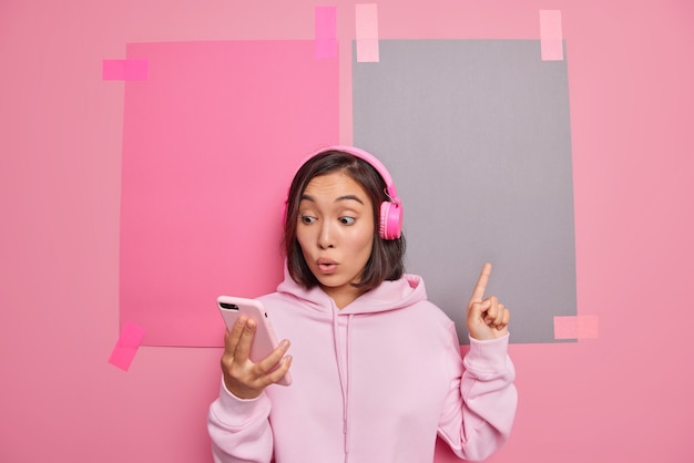 Prise de vue en studio d'une fille asiatique impressionnée montre un espace de copie pour la publicité regarde l'écran du smartphone écoute la piste audio montre la promotion sur le dessus vêtue d'un sweat à capuche décontracté isolé sur un mur rose.