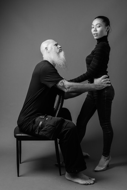 Prise de vue en studio d'un homme chauve barbu mature et d'une belle jeune femme asiatique ensemble contre un mur gris en noir et blanc