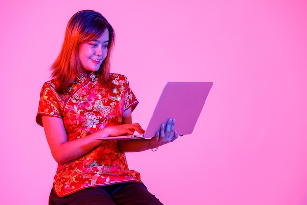 Prise de vue en studio d'une jeune adolescente asiatique millénaire dans une belle chemise qipao cheongsam traditionnelle chinoise rouge assise tenant un ordinateur portable surfant des achats en ligne sur fond rose clair.