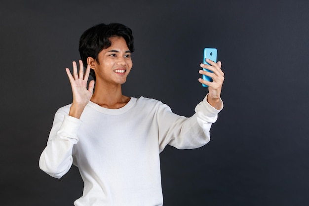 Prise de vue en studio d'un jeune asiatique LGBTQ gay glamour beau modèle masculin homosexuel bisexuel en tenue décontractée debout souriant tenant un appel vidéo sur smartphone saluant dire bonjour salut au revoir sur fond noir