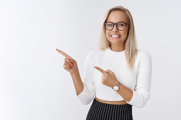 Prise de vue en studio d'une jeune étudiante excitée, heureuse et submergée dans des lunettes élégantes, souriante et enthousiaste en pointant vers le coin supérieur gauche sur une superbe promo sur fond blanc.