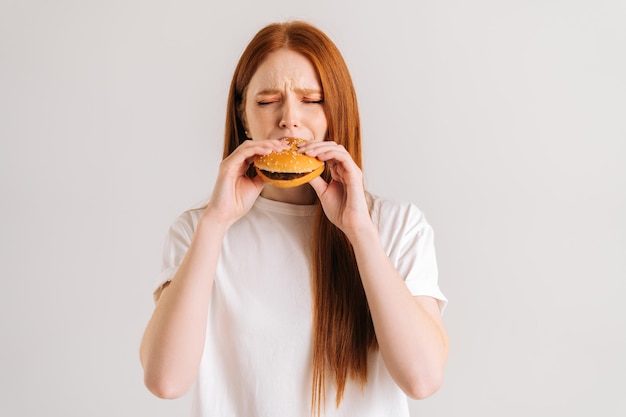 Prise de vue en studio d'une jeune femme affamée heureuse avec les yeux fermés appréciant la bouchée d'un délicieux hamburger appétissant sur fond blanc isolé Vue de face en gros plan d'une femme mangeant un hamburger savoureux