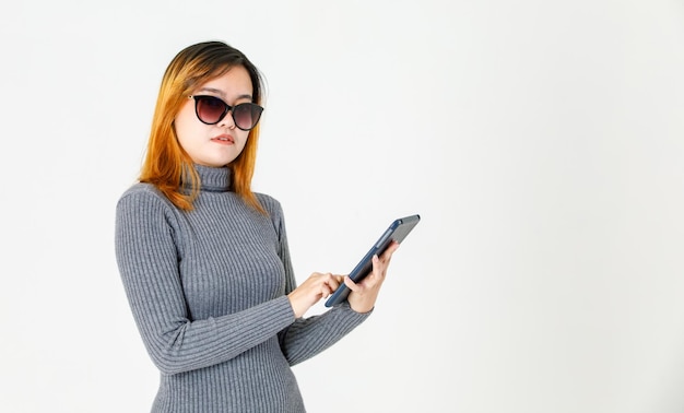 Prise De Vue En Studio D'un Jeune Mannequin Asiatique Aux Cheveux Teints En Pull à Col Roulé Gris Debout Tenant La Main Touchant Des Lunettes De Soleil à Lentille Noire De Mode Tenant Une Tablette Sur Fond Blanc.