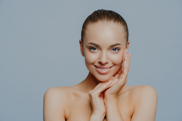 Prise de vue en studio d'une jolie femme souriante touche le visage et jouit d'une peau impeccable, se tient les épaules nues, a un grand front, pose sur fond bleu, applique des cosmétiques de soin de la peau. Tendresse, maquillage