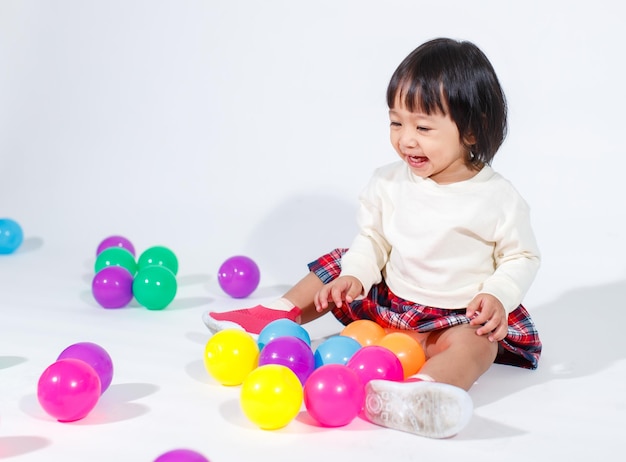 Prise de vue en studio d'un petit modèle asiatique de petite fille aux cheveux noirs courts et mignons en jupe à carreaux décontractée assis sur le sol souriant en riant jouant avec des balles rondes colorées jouet seul sur fond blanc.