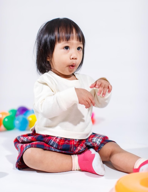 Prise de vue en studio d'un petit modèle asiatique de petite fille aux cheveux noirs courts et mignons en jupe à carreaux décontractée assis sur le sol souriant en riant jouant avec des balles rondes colorées jouet seul sur fond blanc.