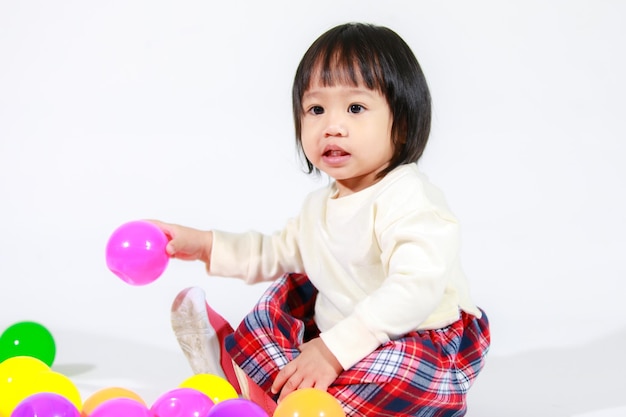 Prise de vue en studio d'un petit modèle asiatique de petite fille aux cheveux noirs courts et mignons en jupe à carreaux décontractée assis sur le sol souriant en riant jouant avec des balles rondes colorées jouet seul sur fond blanc.