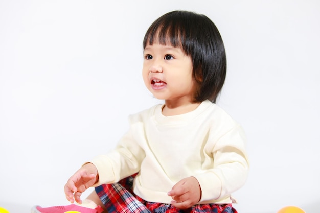 Prise de vue en studio d'un petit modèle asiatique de petite fille aux cheveux noirs courts et mignons en jupe à carreaux décontractée assis sur le sol souriant en riant jouant avec des balles rondes colorées jouet seul sur fond blanc.