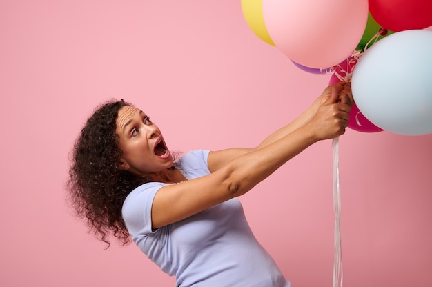 Photo prise de vue en studio pour la publicité d'une jolie femme surprise qui cherche des ballons à air colorés, debout sur fond rose avec espace de copie. le concept de célébration, de vacances, d'anniversaire et de cadeaux.