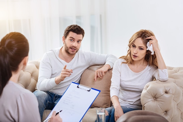 Problèmes dans les relations. Beau jeune homme sans joie assis à côté de sa femme et parlant au psychologue de leur problème familial tout en ayant une séance psychologique