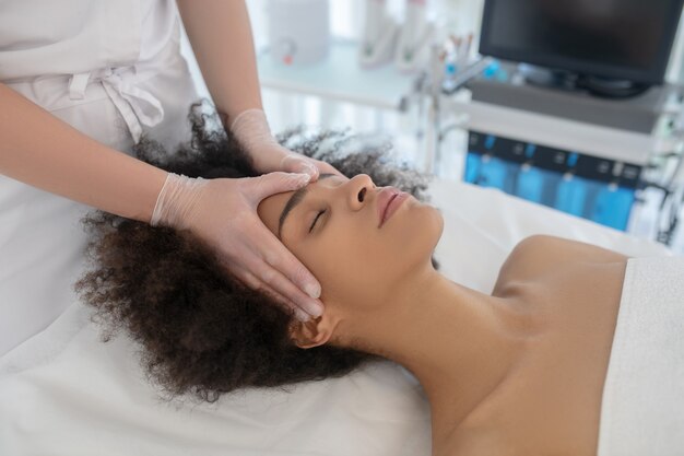 Photo procédure cosmétologique. mains de l'esthéticienne dans les gants sur le visage d'une fille mulâtre allongée sur le canapé