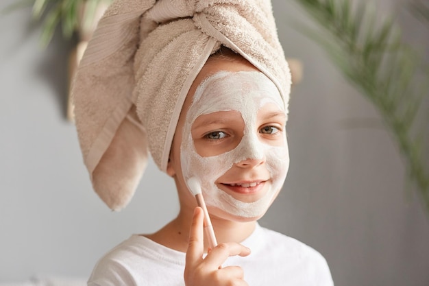 Procédures de spa facial à la maison Amusante petite fille mignonne dans une serviette de bain blanche avec un masque cosmétique sur le visage regardant la caméra avec une expression joyeuse bénéficiant d'un traitement de soins de la peau