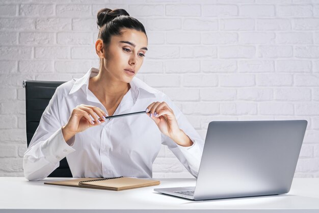 Processus d'affaires et concept de travail avec une jolie jeune femme assise au bureau blanc regardant un écran d'ordinateur portable et portant un crayon dans les mains sur fond de mur de briques claires