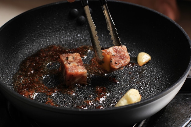 Processus de cuisson, steak de boeuf en cubes de Wagyu Fatty Saikoro grillé avec sauce barbecue sur une poêle noire à l'aide de pinces en acier inoxydable. Steak Epice avec Poivre Noir et Sel