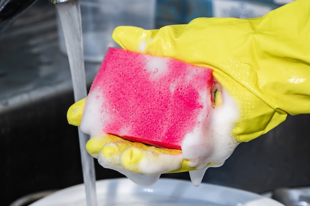 Le processus de lavage de la vaisselle sale. Une femme dans un gant jaune tient une éponge rose avec de la mousse.
