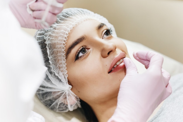 Processus de préparation pour l'amélioration des lèvres. Le médecin aspire de l'acide hyaluronique devant le patient. La jeune fille avec un beau visage dans le chapeau spécial et les mains du médecin en gant rose