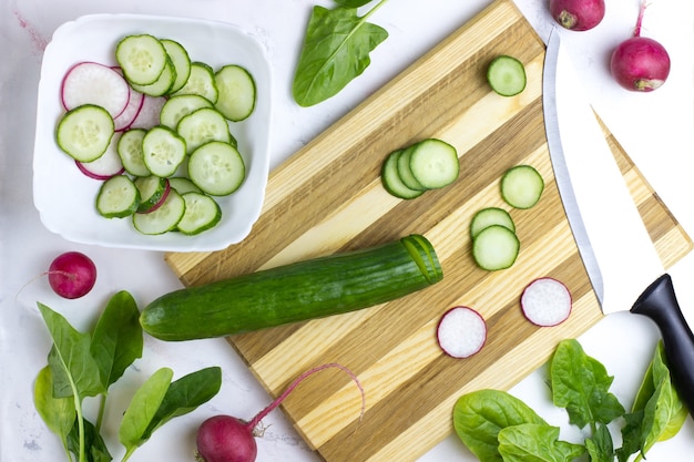 Le processus de préparation de la salade. Couper le concombre sur une planche de bois. Salade de concombre, radis et épinards. Vue de côté