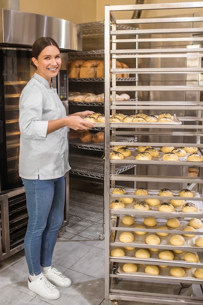 Processus de préparation. Sourire jeune femme adulte debout près du four et de la grille avec des plateaux de petits pains en boulangerie