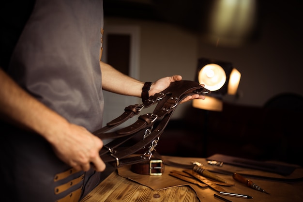 Processus de travail de la ceinture en cuir dans l'atelier de cuir. Homme tenant la ceinture du photographe pour appareil photo. Outil sur fond en bois. Tanner dans l'ancienne tannerie. Bouchent le bras du maître