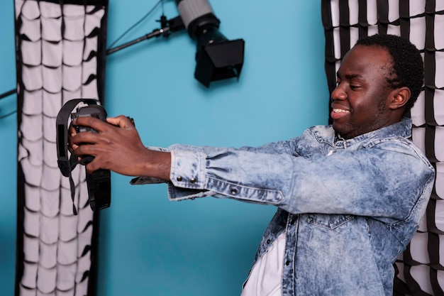 Producteur de studio professionnel prenant une photo de selfie avec un appareil photo reflex numérique tout en se tenant à côté d'un équipement photo moderne sur fond bleu. Jeune photographe prenant une photo de lui-même en souriant chaleureusement.