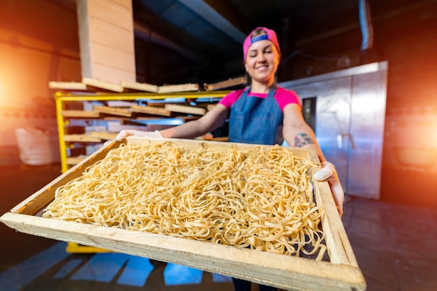 Production de pâtes à l'usine. Boîtes en bois avec des nouilles dans les mains du boulanger. Travail industriel d'usine de production technologique, gros plan de macaroni cru.