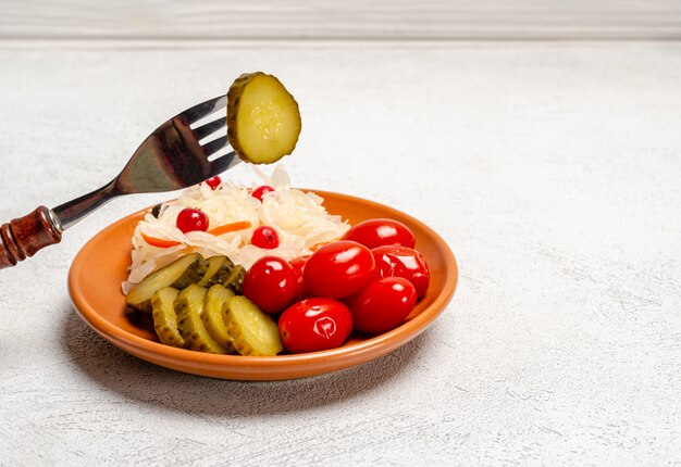 Produits fermentés faits maison sur une assiette, choucroute, tomates marinées, cornichons, fond gris clair. Sur une fourchette est une tranche de concombre mariné.