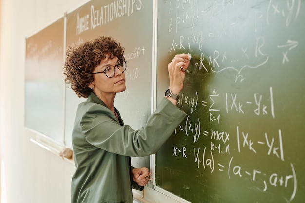 Photo professeur féminin sérieux d'algèbre écrivant des équations sur le tableau noir