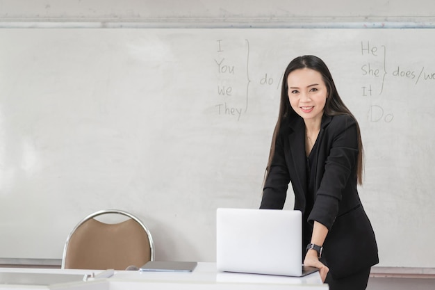 Professeur de femme asiatique gaie et confiante adulte en uniforme de costume noir avec tablette numérique et ordinateur portable enseignant moderne en classe