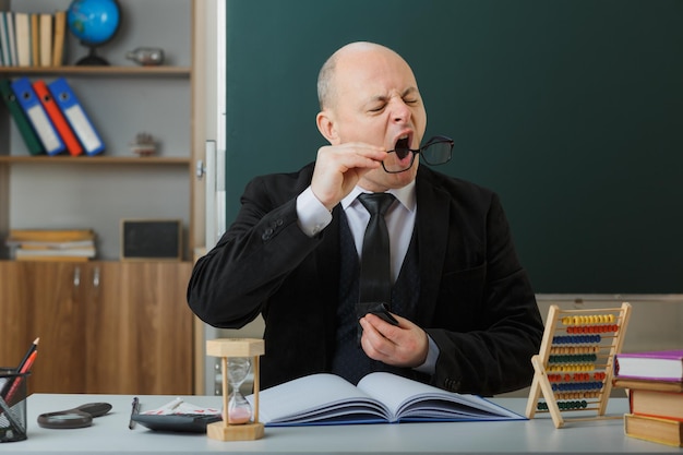 Professeur d'homme vérifiant le registre de classe en essuyant ses lunettes assis au bureau de l'école devant le tableau noir dans la salle de classe