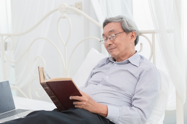 Professeur de lunettes de vieil homme asiatique aiment lire le livre de texte tous les matins sur le lit