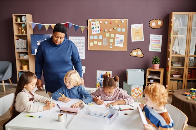 Professeur de pépinière afro-américain mature debout près de la table où les enfants dessinent