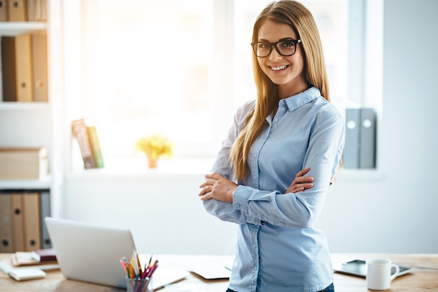 Professionnel et beau. Joyeuse jeune femme à lunettes en gardant les bras croisés et en regardant la caméra avec le sourire en se tenant debout sur son lieu de travail