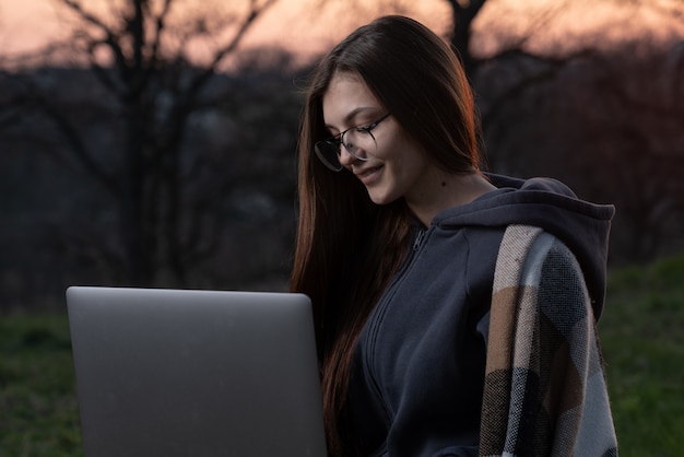 Professionnel indépendant pensif travaillant sur un ordinateur portable et réfléchissant à un projet à l'extérieur. Belle jeune femme à lunettes ditting sur couverture et herbe et à l'aide d'ordinateur.