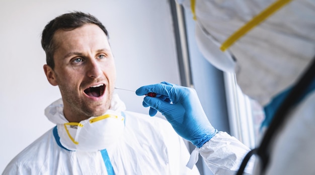 Un professionnel de la santé en vêtements de protection prend un tube à essai sur écouvillon de la bouche d'un clinicien épuisé dans un centre de test Covid19 pendant le processus de protocole de test ADN PCR de l'épidémie de coronavirus