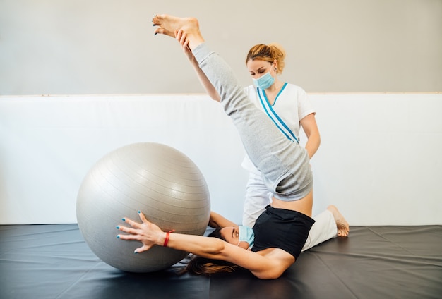 Professionnelle De Physiothérapie De Race Blanche Dans Une Clinique Donnant Un Traitement De Pilates Avec Une Balle De Thérapie à Un Client Avec Un Masque Facial En Raison De La Pandémie De Coronavirus Covid 19.