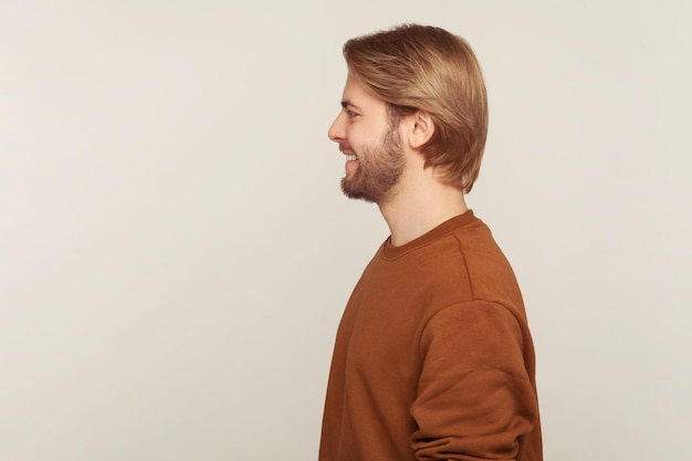 Profil d'un bel homme joyeux et joyeux avec des cheveux et une barbe soignés portant un sweat-shirt debout, souriant à côté