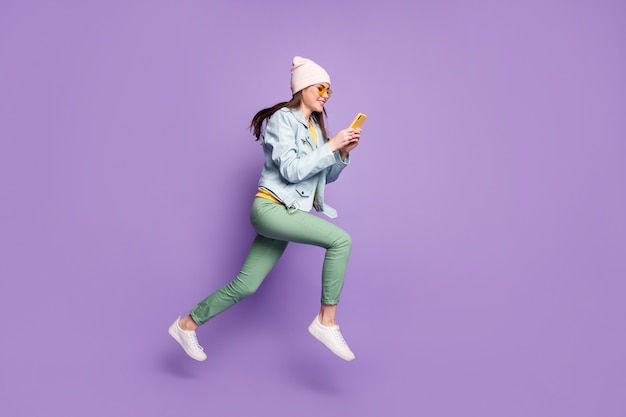 Profil complet du corps photo de côté fille positive courir utiliser un smartphone suivre les médias sociaux republier après porter une veste en cuir pantalon vert pantalon lunettes de soleil jaune isolé fond de couleur violet