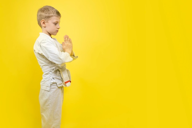 Profil complet d'un garçon en kimono de karaté isolé sur fond jaune Enfant tient les mains ensemble dans la salutation traditionnelle Garçon de karaté avec un visage concentré en uniforme Bannière publicité de karaté