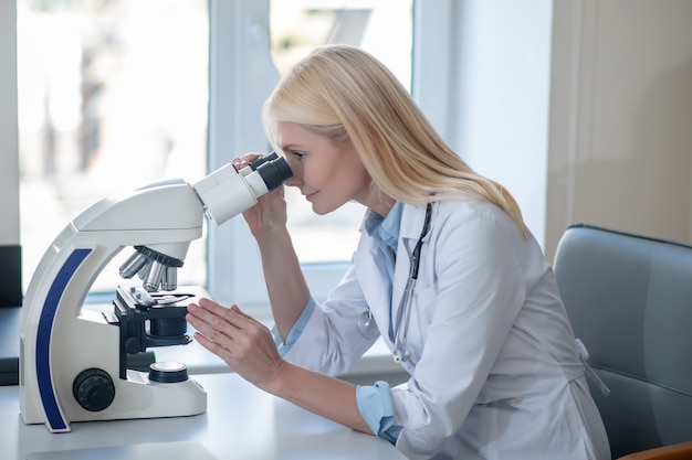 Profil de femme aux cheveux longs en blouse blanche à la recherche au microscope assis dans une pièce lumineuse