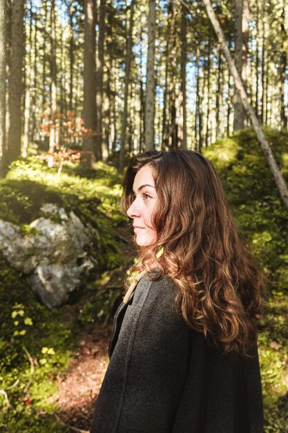 Profil d'une jeune femme aux cheveux bruns dans un long manteau marche seule au milieu d'une forêt dense