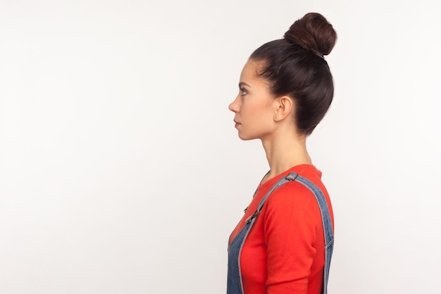 Photo profil d'une jolie fille mince et élégante avec un chignon dans une salopette en denim debout avec un visage sérieux, une expression pensive calme, regardant l'espace de copie latéral. studio d'intérieur tourné isolé sur fond blanc