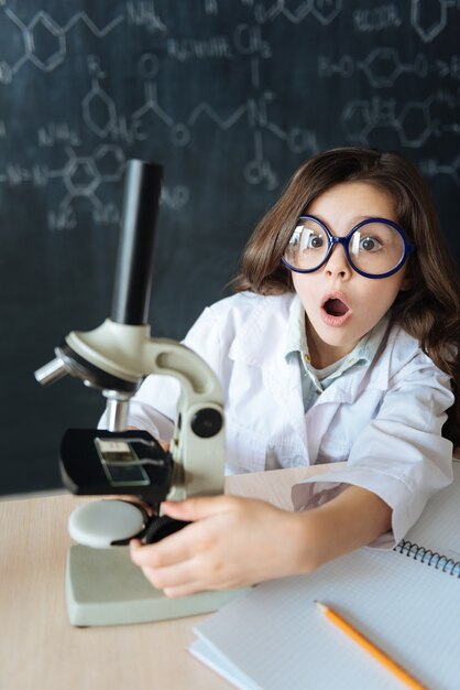 Profitant D'une Invention écrasante. étonné, Stupéfait, Jeune Fille Assise Dans Le Laboratoire Et Bénéficiant D'un Cours De Chimie Tout En Participant à L'expérience Scientifique Et En Utilisant Un Microscope