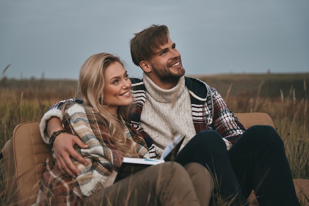 Profitant de leur liberté. Beau jeune couple embrassant et regardant ailleurs avec le sourire alors qu'il était assis sur le terrain