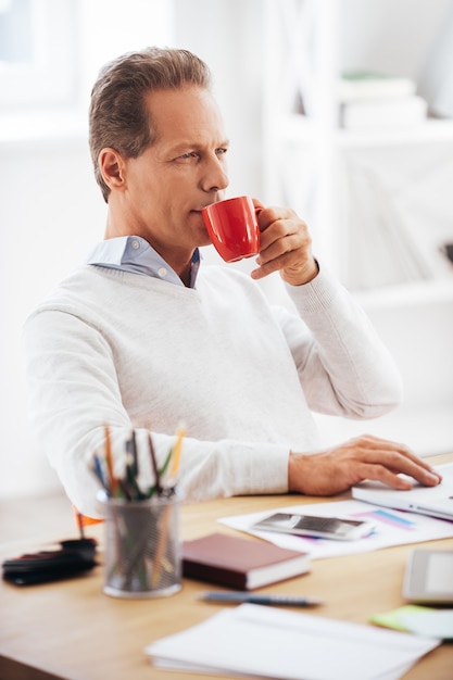 Profitant de sa pause-café Homme mûr confiant buvant du café et regardant ailleurs alors qu'il était assis sur son lieu de travail