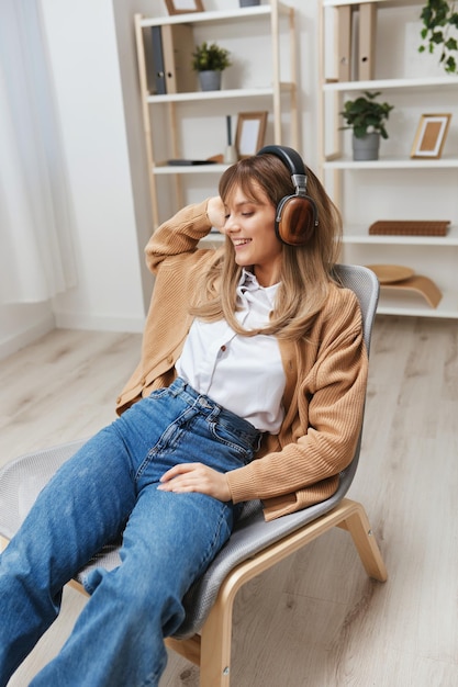 Profité du temps libre jeune étudiante blonde pigiste en pull chaud dans les écouteurs écouter des chansons fav assis dans un fauteuil à l'intérieur de la maison moderne Temps de musique Relaxing Cool playlist Concept Copy space