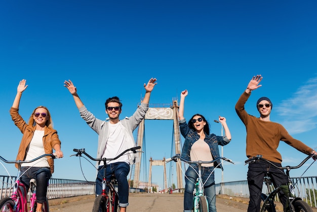 Profiter d'une balade amusante. Vue en contre-plongée de quatre jeunes gens joyeux faisant du vélo et gardant les bras levés